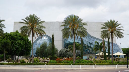 Moderne Museumsfassade mit einem großen rechteckigen Betonbau, aus dem eine organisch geformte Glaskuppel aus dreieckigen Scheiben hervortritt. Davor liegt ein angelegter Garten mit Palmen, Sträuchern, Felsen und roten Blumen. Der Himmel ist hell und leicht bewölkt.