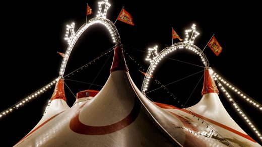 Weiß-rotes Zirkuszelt, dessen Spitzen in den dunklen Nachhimmel ragen und durch Leuchtbögen miteinander verbunden sind.